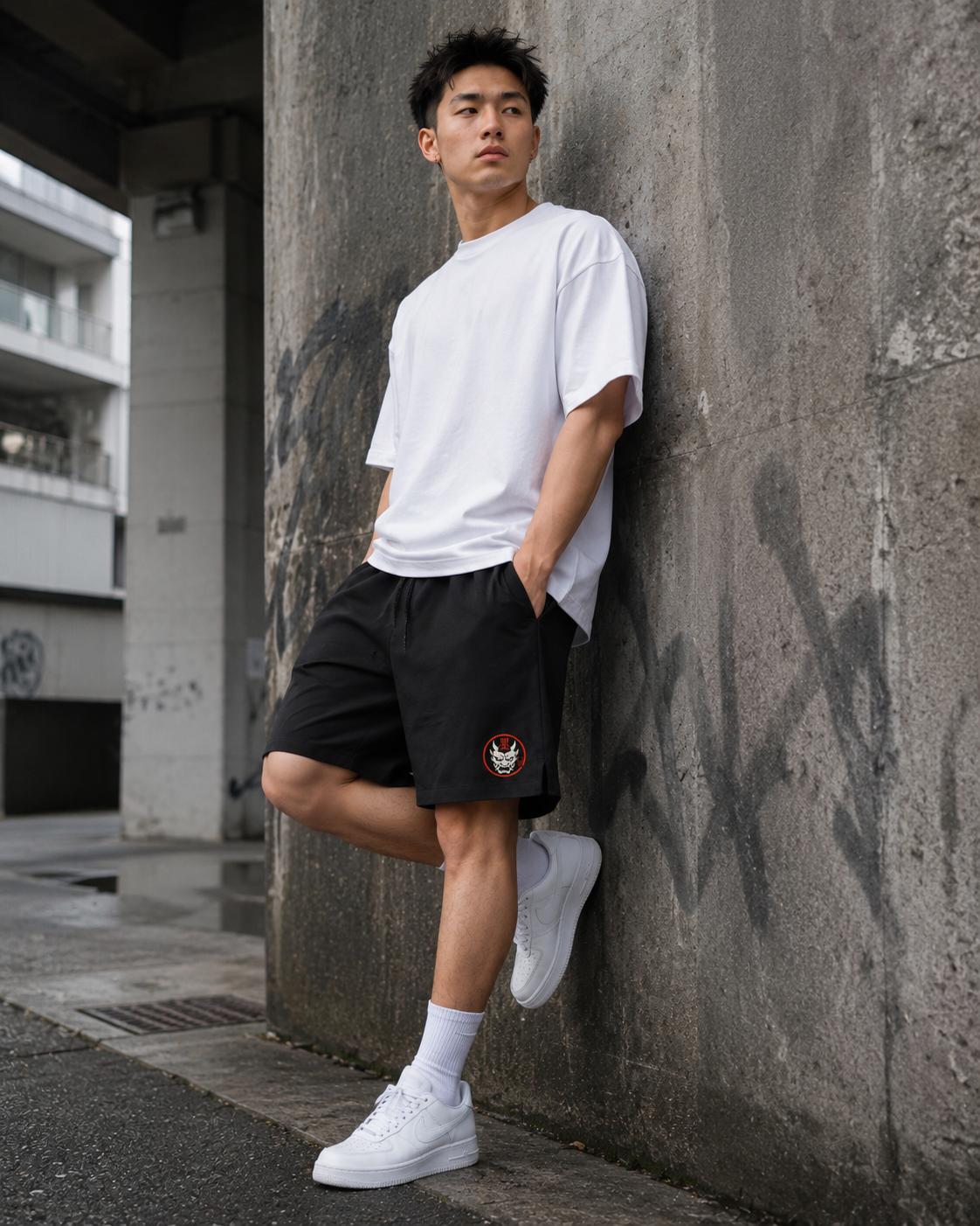 A young man in a white T-shirt, Kuro Oni Streetwear Shorts with a red and white patch, white socks, and white trainers leans against a graffiti-covered concrete wall in an urban setting.