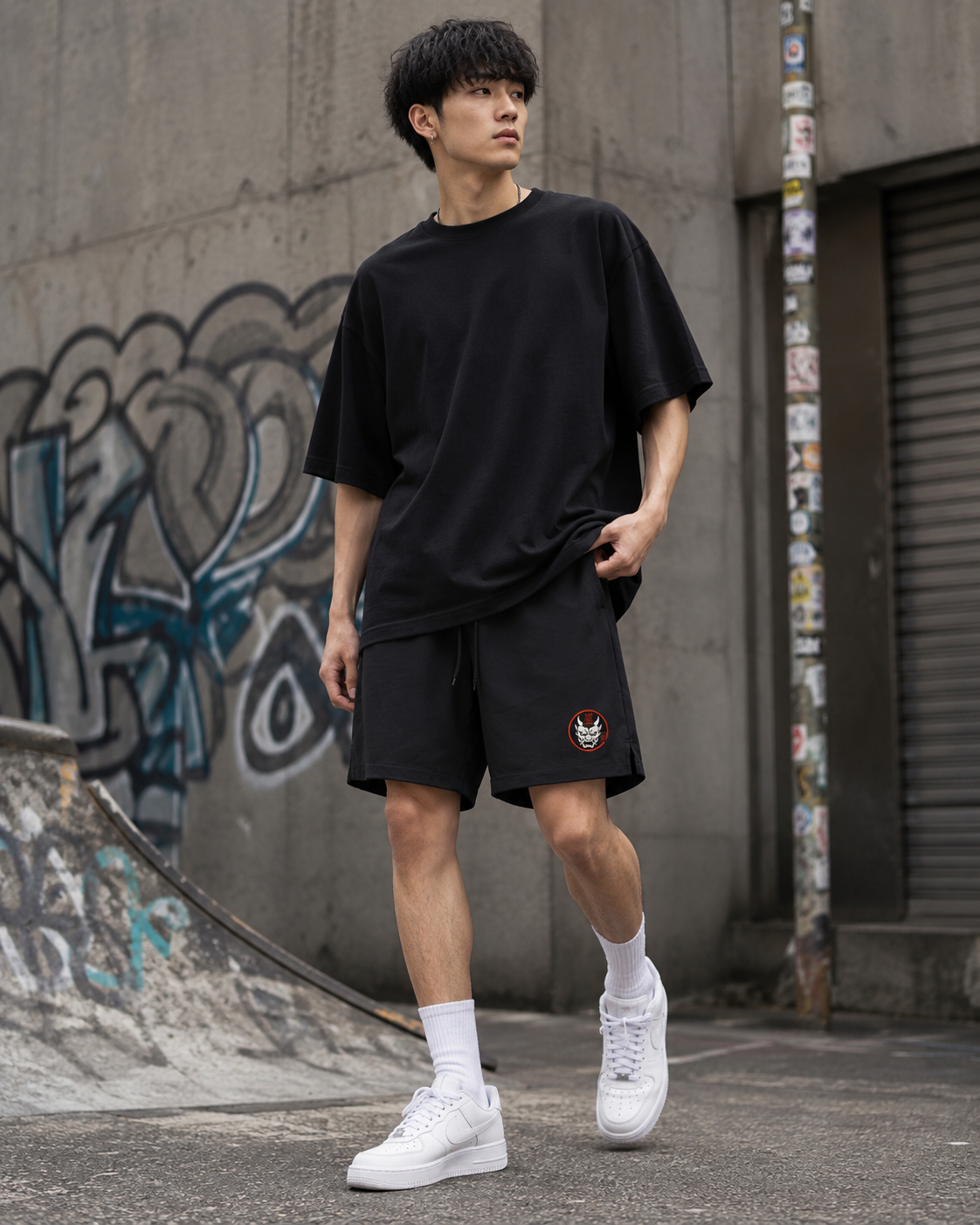 A young man wearing a loose black Irezumi Ink Clothing T-shirt, Kuro Oni Streetwear Shorts, white socks, and white trainers walks in an urban skate park with graffiti-covered walls.